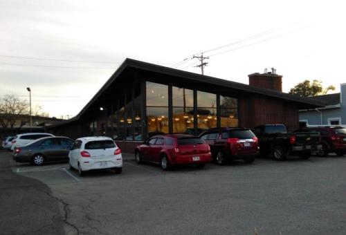 A modern building with large windows, surrounded by parked cars in a lot during dusk.