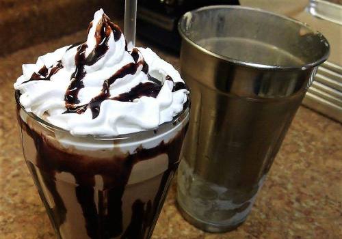 A tall glass filled with a chocolate dessert topped with whipped cream and chocolate drizzle, next to a metal cup.