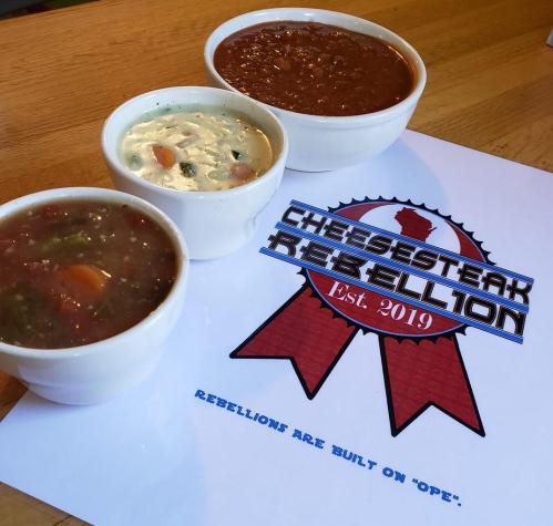 Three bowls of soup on a wooden table, with a logo for "Cheesesteak Rebellion" and the text "Rebellions are built on 'Ope'."