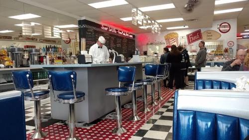 A retro diner interior with blue stools, a counter, and a chef preparing food, featuring a checkered floor and colorful decor.