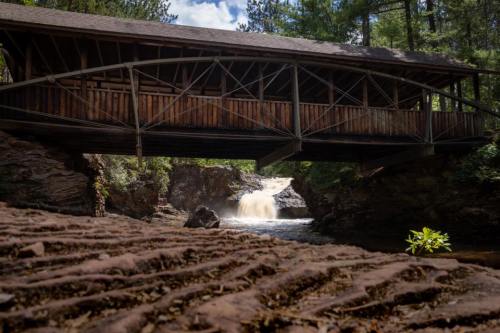 A wooden covered bridge spans a rocky river with a waterfall, surrounded by lush greenery and blue skies.