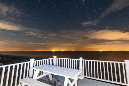 A white picnic table on a deck overlooking a starry night sky and calm ocean waters with distant lights on the horizon.
