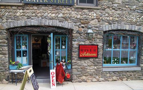 A stone building with blue windows and an "Open" sign, featuring a red sign for "Connecticut River Artisans."