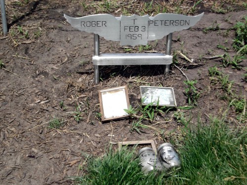 A metal grave marker with the name "Roger T. Peterson" and the date "Feb 3, 1959," surrounded by framed photos and jars.