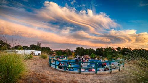 A scenic view of a circular gathering area with people, surrounded by greenery and vibrant clouds at sunset.