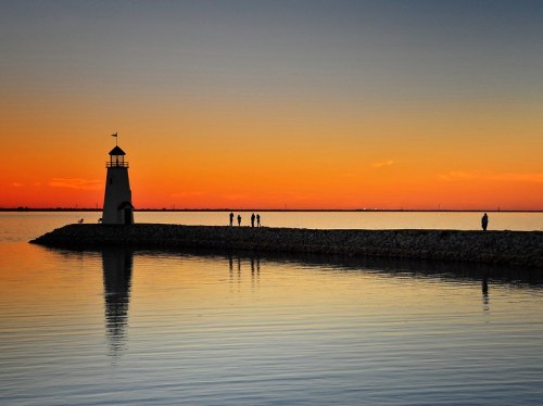 Silhouette of a lighthouse at sunset, with people walking along a pier and calm water reflecting the colorful sky.