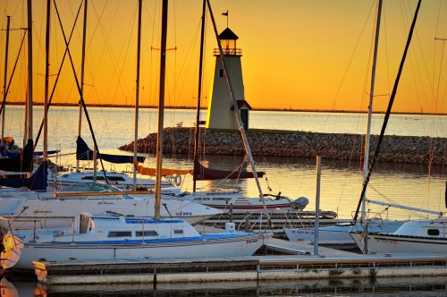 Sailboats docked at a marina with a lighthouse at sunset, casting warm reflections on the water.