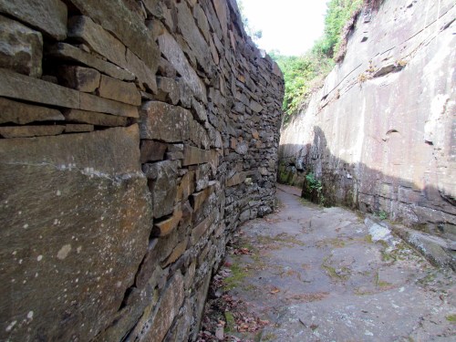 A narrow stone pathway surrounded by tall, weathered rock walls, with greenery peeking through the cracks.
