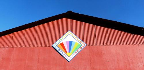 A colorful diamond-shaped mural on a red barn against a clear blue sky.