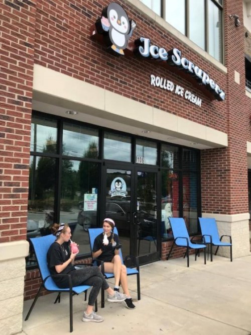 Two people enjoy rolled ice cream outside Ice Scraperz, a shop with a penguin logo, on a sunny day.