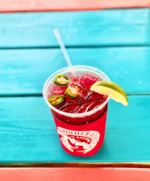 A refreshing red drink with lime and jalapeño slices in a clear cup on a colorful wooden table.