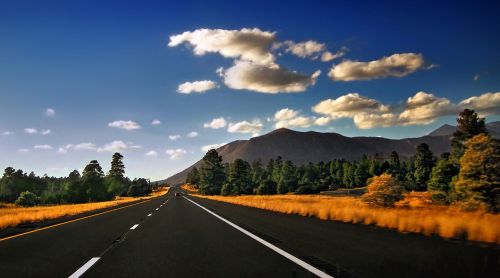 A scenic highway stretches through golden fields and green trees, with mountains and a blue sky in the background.