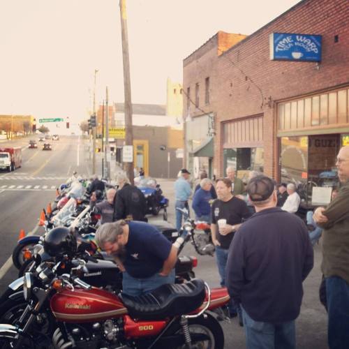 A group of people gathered near parked motorcycles outside a shop on a street during an event.