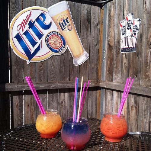 Three colorful cocktails in round glasses with pink straws, set against a wooden backdrop featuring Miller Lite signs.
