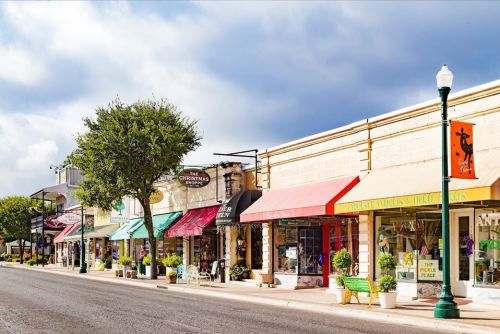 Charming street lined with shops featuring colorful awnings and greenery under a partly cloudy sky.