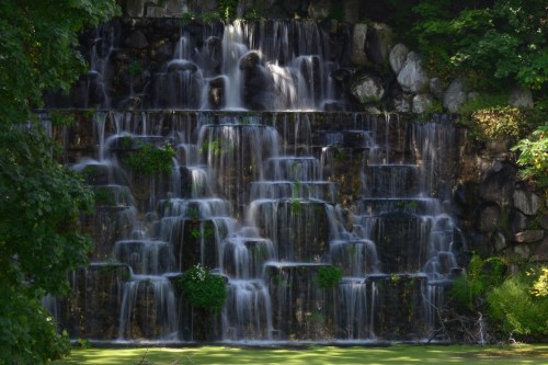 A cascading waterfall flows over rocky tiers, surrounded by lush greenery and a tranquil pond below.