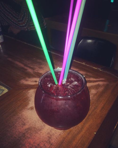 A colorful drink in a large glass with three glowing straws, sitting on a wooden table.