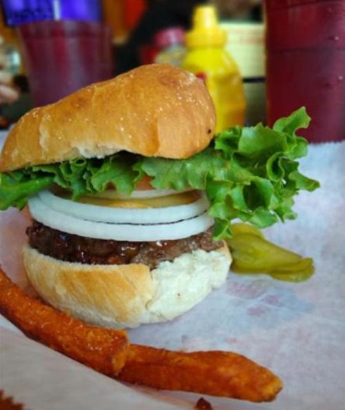A close-up of a hamburger with lettuce, tomato, onion, and pickles, served with a side of sweet potato fries.