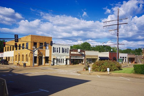 A small town street with brick buildings, shops, and a clear blue sky with scattered clouds. Power lines in the background.