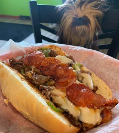 A loaded sandwich with meat, peppers, and cheese, with a small dog peeking in the background.