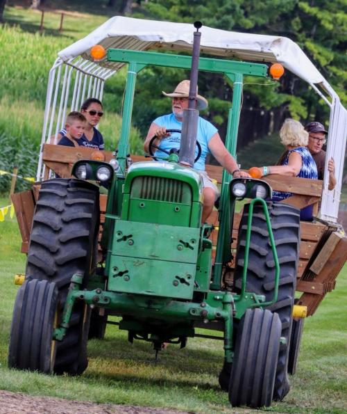 A green tractor pulls a wagon with people enjoying a ride through a scenic field.