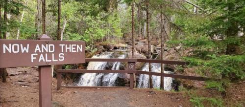 A wooden sign reads "Now and Then Falls" near a serene waterfall surrounded by lush green trees.
