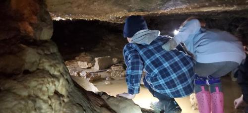 Two children explore a cave, crouching near water with headlamps illuminating the rocky surroundings.