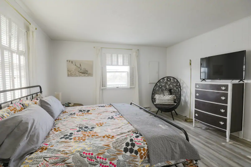 Cozy bedroom with a patterned bedspread, a TV, a decorative chair, and natural light from a window.