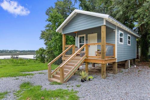A small blue house on stilts with a wooden porch, surrounded by greenery and near a body of water.
