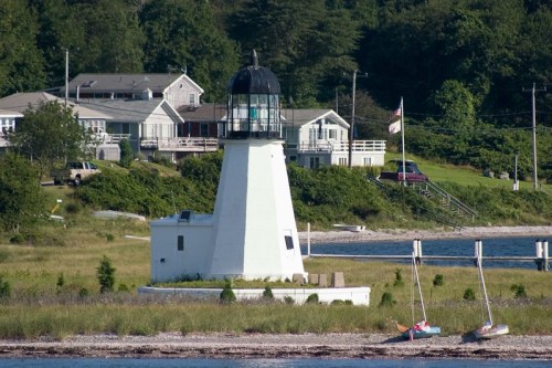 A white lighthouse with a black lantern sits near a grassy shore, surrounded by trees and a few boats in the water.