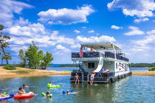 A houseboat on a lake with people swimming and relaxing on inflatable floats under a bright blue sky.