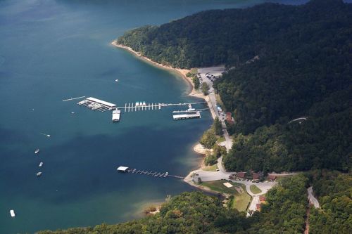 Aerial view of a marina on a lake, surrounded by green hills and a winding road along the shore.