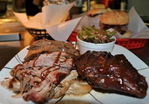 A plate of barbecue meats with sauce, served alongside green beans and a sandwich in the background.