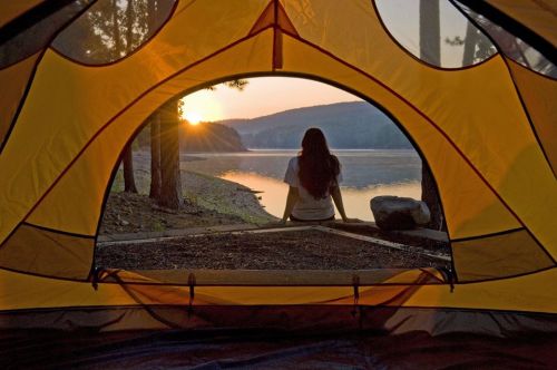 A person sits inside a tent, gazing at a sunset over a calm lake surrounded by mountains.