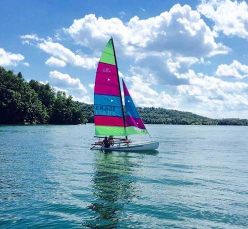 A colorful sailboat with a vibrant sail glides across a calm lake under a blue sky with fluffy clouds.