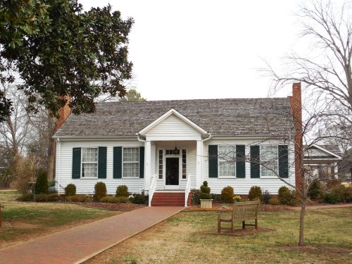A charming white house with green shutters, a front porch, and a brick pathway, surrounded by neatly trimmed shrubs.
