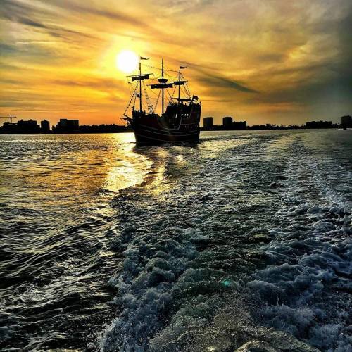 A silhouette of a pirate ship against a vibrant sunset, with waves trailing behind it.