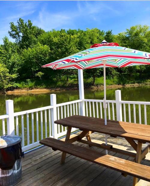 A wooden picnic table with a colorful striped umbrella by a calm river, surrounded by lush green trees.