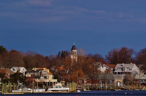 A scenic view of a waterfront town with a clock tower, boats, and trees under a cloudy sky.
