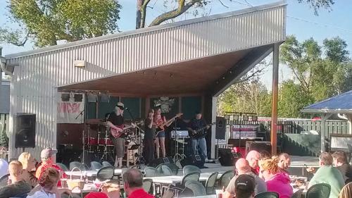 A live band performs on an outdoor stage, with an audience seated at tables in a sunny setting.