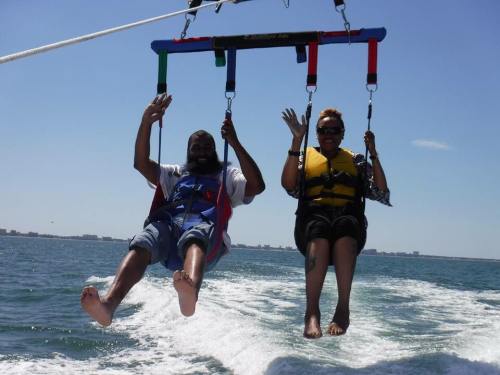 Two people are parasailing over the ocean, smiling and waving, with a clear blue sky in the background.