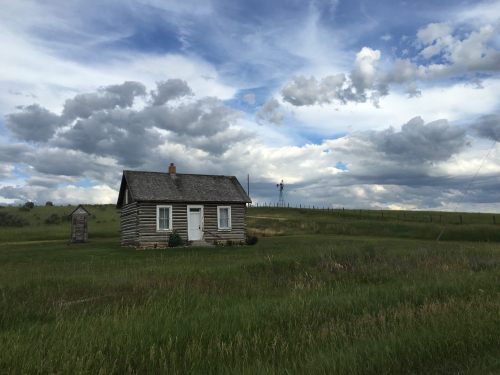 A rustic log cabin sits in a grassy field under a dramatic sky filled with clouds.