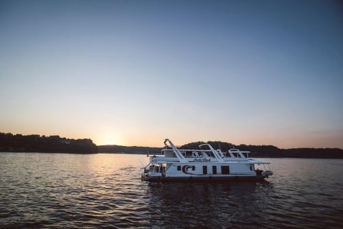 A houseboat floats on calm water at sunset, with a serene landscape in the background.
