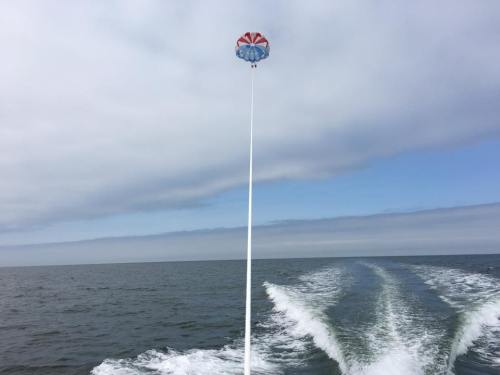 A colorful parachute is soaring high above the ocean, trailing behind a boat on a cloudy day.