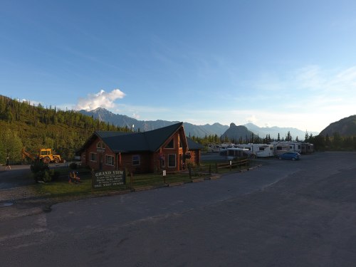 A log cabin with a green roof, surrounded by mountains and RVs, under a clear blue sky. Sign reads "Grand View."
