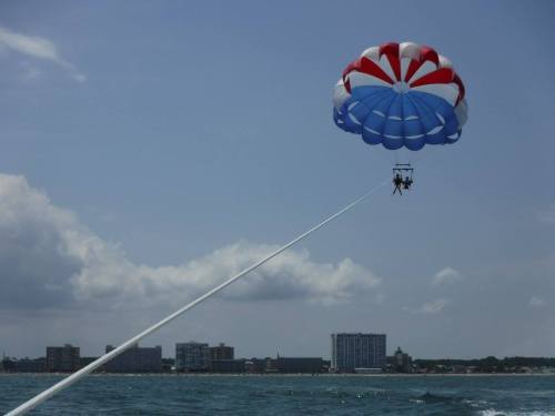 Two people parasailing high above the ocean, with a colorful parachute and a coastal city skyline in the background.