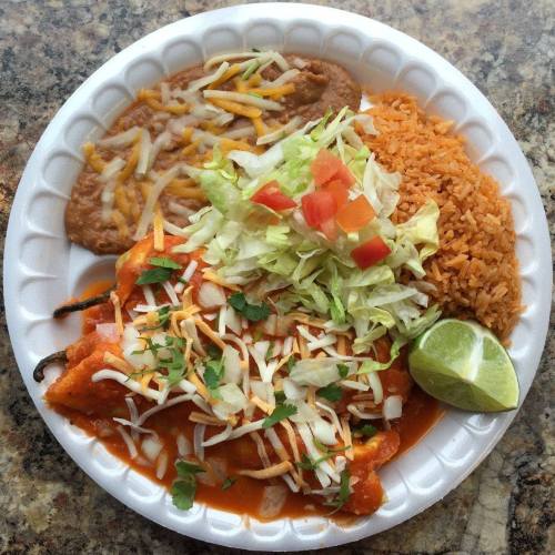 A plate of enchiladas topped with cheese and sauce, served with rice, refried beans, lettuce, tomatoes, and a lime wedge.
