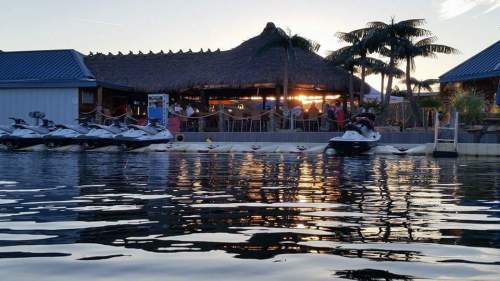 A waterfront scene at sunset featuring jet skis, a thatched-roof bar, and palm trees reflecting on the water.