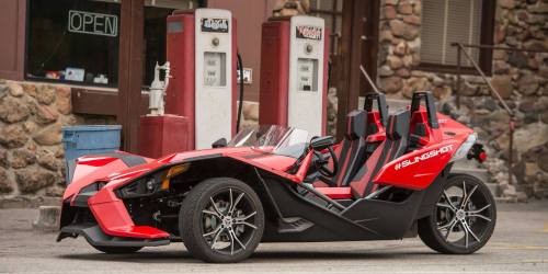 A red three-wheeled vehicle parked near vintage gas pumps in a rustic setting.