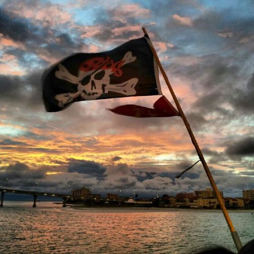 A pirate flag waves against a colorful sunset over a river, with buildings and a bridge in the background.